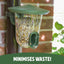 Green bird feeder filled with seeds attached to a brick wall with text 'MINIMISES WASTE!' at the bottom.