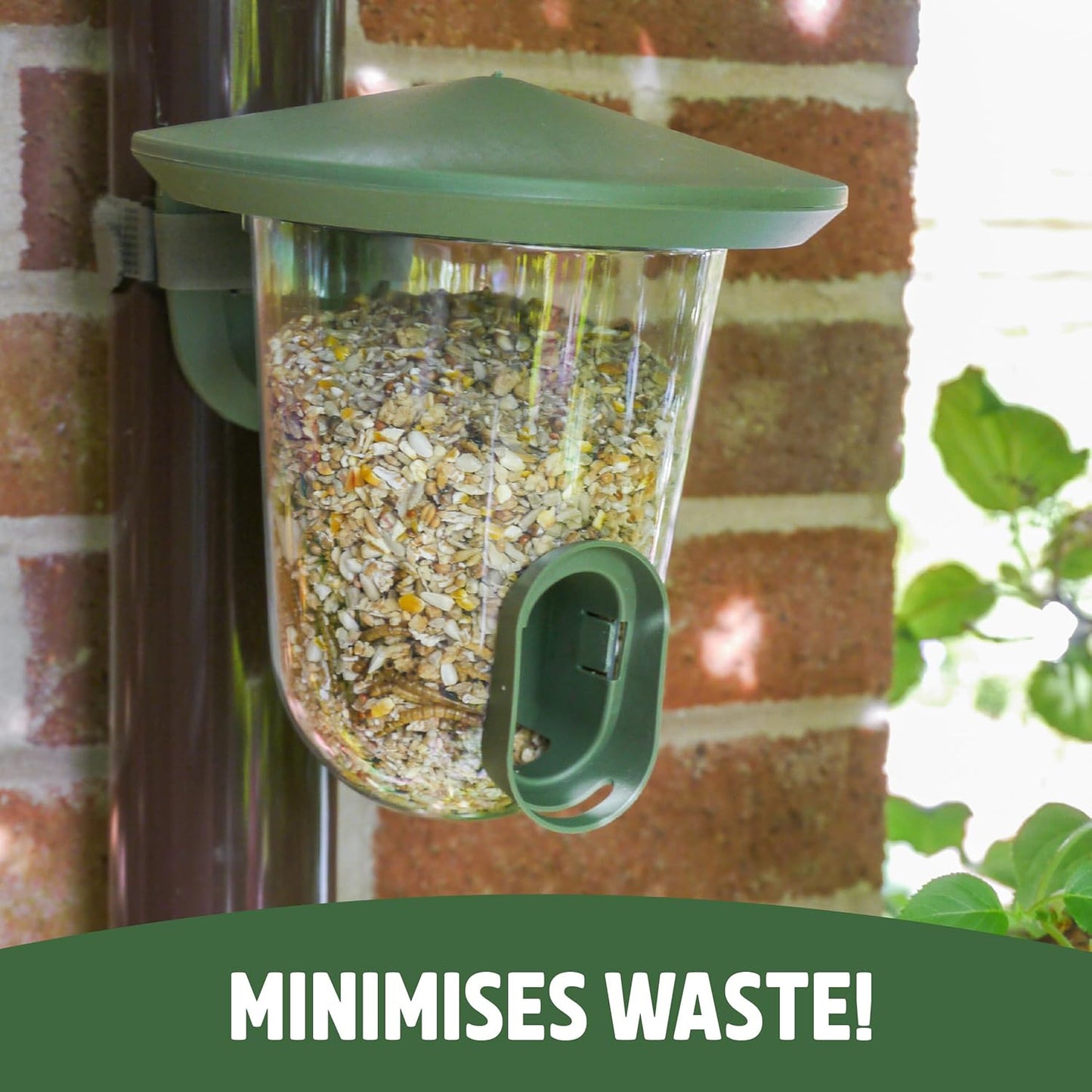 Green bird feeder filled with seeds attached to a brick wall with text 'MINIMISES WASTE!' at the bottom.