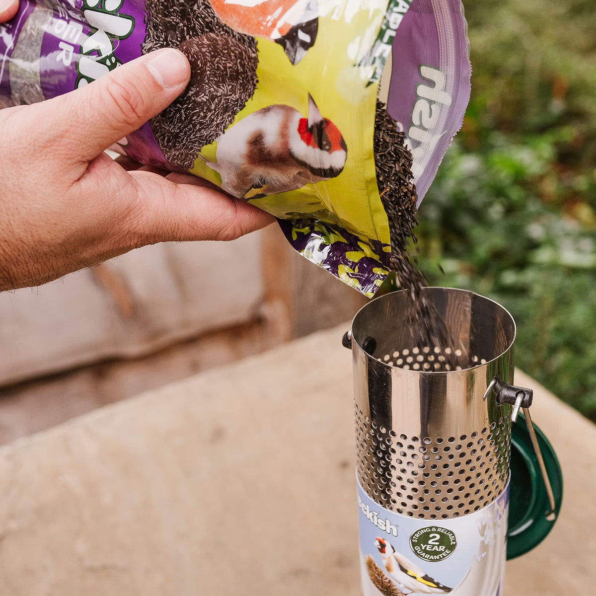 A hand pours birdseed from a colorful bag into a stainless steel percolator outdoors on a table, with a blurred garden background.