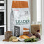 A bag of Leader Natural Dog Nutrition adult dog food stands on a countertop, surrounded by ingredients including kibble in a glass jar, dried herbs, a piece of chicken, beets, and greens. A second glass jar containing the dry food is also visible to the right.
