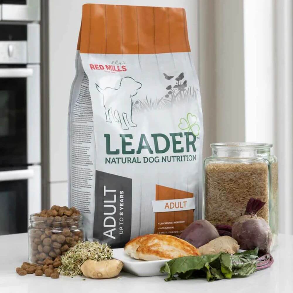 A bag of Leader Natural Dog Nutrition adult dog food stands on a countertop, surrounded by ingredients including kibble in a glass jar, dried herbs, a piece of chicken, beets, and greens. A second glass jar containing the dry food is also visible to the right.