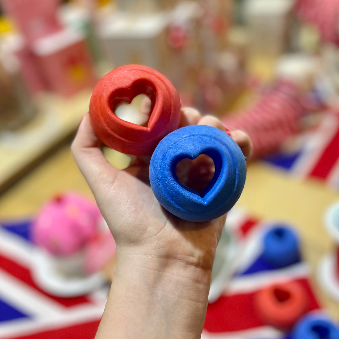 Hand holding two heart-shaped balls, one red and one blue, with a blurred background of similar items.