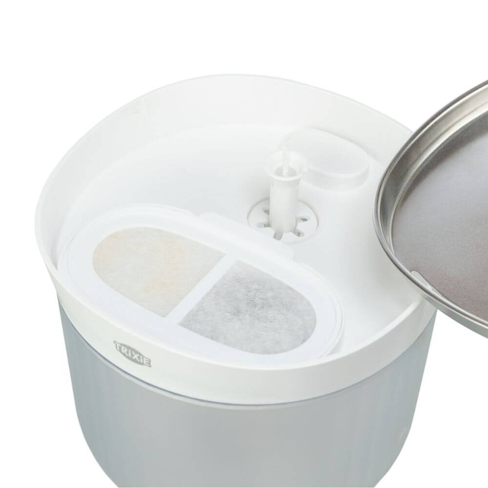 A close-up of a white and light gray pet waterer or feeder container with a stainless steel lid partially visible on the right. The top has a central white spout and two oval filter compartments, one showing a beige filter and the other a gray filter. It is a dog water bowl with fountain, stainless steel bowl, filters and clear container. 